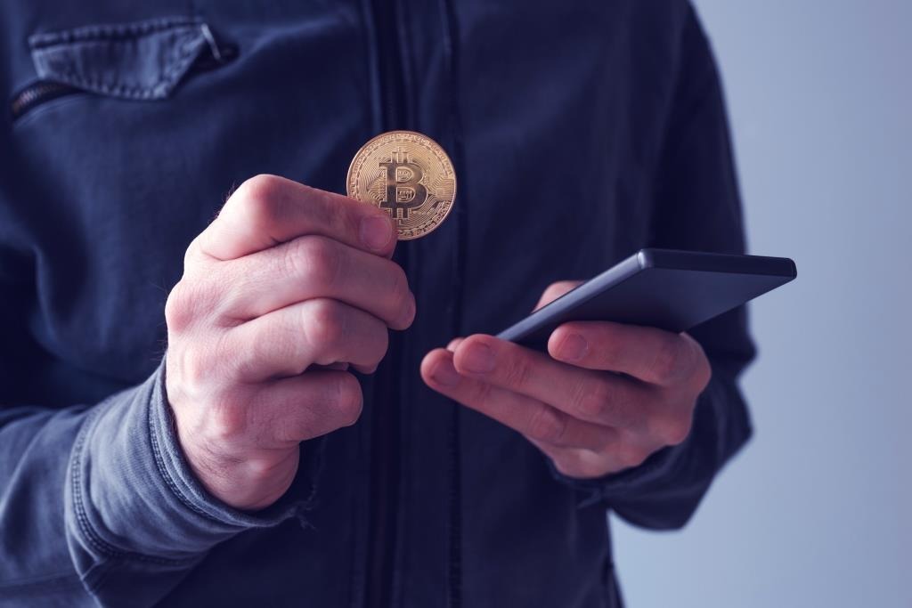 Man in donkerblauwe jas met een gouden bitcoinmunt en een smartphone, beeld bij de fiscaliteit van digitale activa